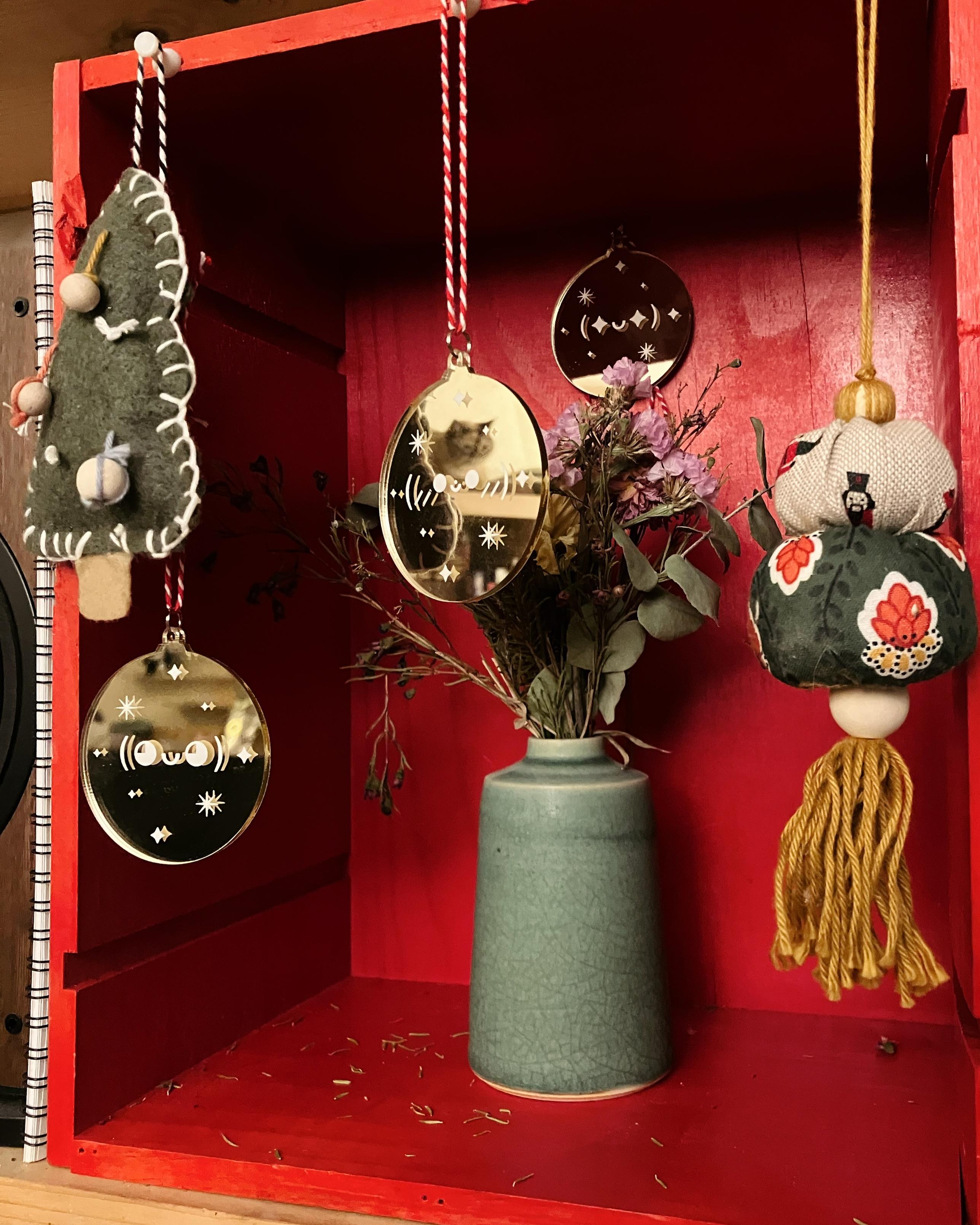 Photo d’une caisse en bois rouge, à l’intérieur de laquelle il y a un bouquet de fleurs séchées, trois décorations rondes effet miroir avec des visages expressifs en caractères typographiques, deux décorations cousues en tissus et feutrine. Dans le reflet d’une des trois décorations on voit la tête d’un chat flou 